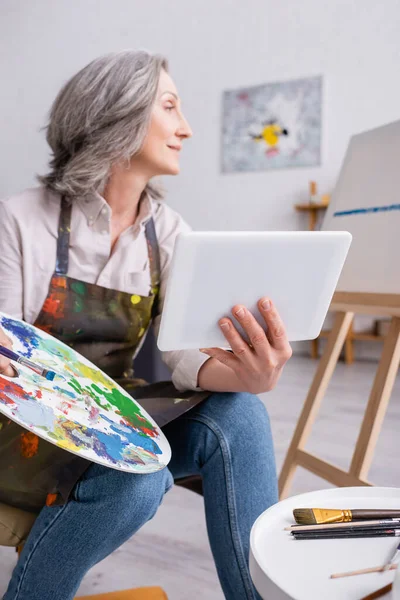 Reife Frau mit Palette und digitalem Tablet beim Betrachten der Leinwand — Stockfoto