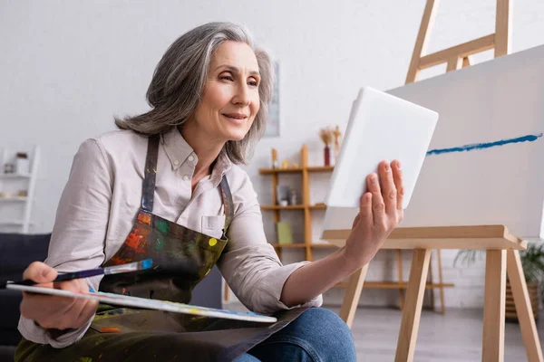 Mature woman holding palette and digital tablet while watching tutorial near canvas — Stock Photo