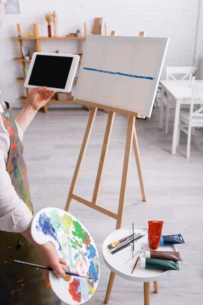 Vista recortada de mujer madura sosteniendo paleta y tableta digital con pantalla en blanco cerca de lienzo - foto de stock