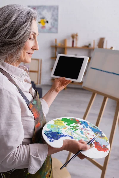Mature woman holding palette, paintbrush and digital tablet with blank screen near canvas on blurred background — Stock Photo