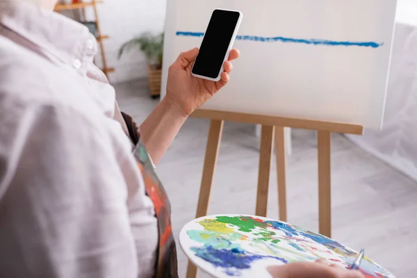 Partial view of mature woman holding palette, paintbrush and smartphone with blank screen near canvas on blurred background — Stock Photo
