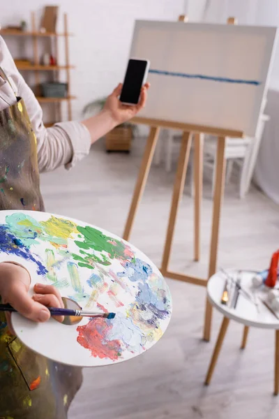 Ausgeschnittene Ansicht einer reifen Frau mit Palette, Pinsel und Smartphone mit leerem Bildschirm nahe der Leinwand auf verschwommenem Hintergrund — Stockfoto