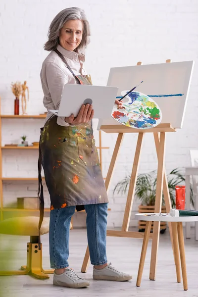 Felice donna matura in possesso di tavolozza e laptop mentre si guarda tutorial vicino tela — Foto stock