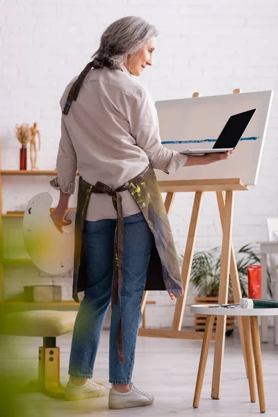 Mujer madura sosteniendo paleta y portátil con pantalla en blanco mientras mira tutorial cerca de lienzo - foto de stock