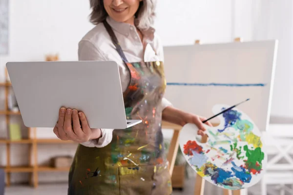 Vista recortada de la mujer madura sosteniendo la paleta y el ordenador portátil mientras mira tutorial cerca de lienzo — Stock Photo