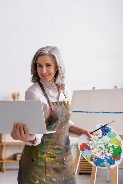 Smiling mature woman holding palette and laptop while watching tutorial near canvas — Stock Photo