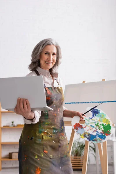 Joyeuse femme mature tenant palette et ordinateur portable près de la toile — Photo de stock