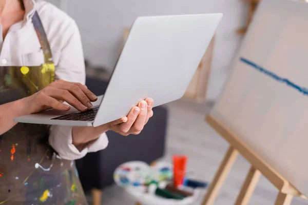 Partial view of mature woman holding laptop while watching tutorial near canvas — Stock Photo