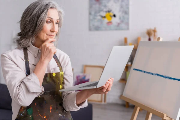 Mujer madura sosteniendo portátil mientras mira tutorial cerca de lienzo - foto de stock