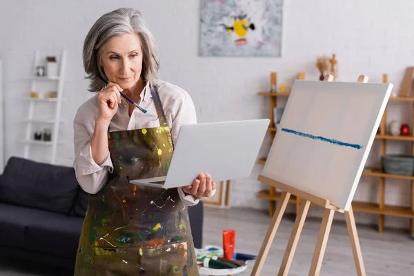 Donna matura che tiene pennello e laptop mentre guarda tutorial vicino alla tela — Foto stock