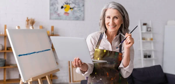 Happy mature artist holding paintbrush and laptop near canvas, banner — Stock Photo