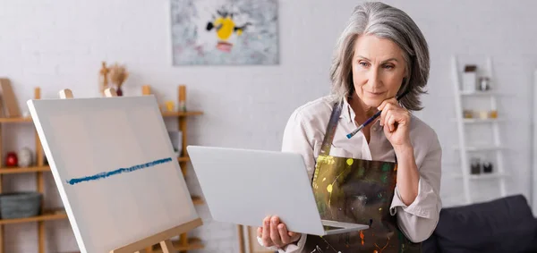 Reife Frau mit Pinsel und Laptop, während sie Tutorial in der Nähe von Leinwand, Banner — Stockfoto