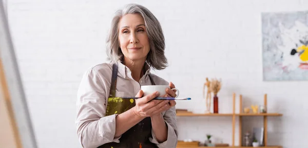 Artista de mediana edad sosteniendo taza de café y pincel cerca de la lona, pancarta - foto de stock