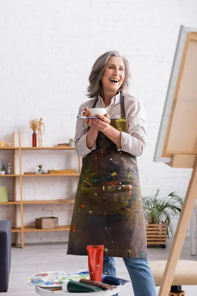 Aufgeregter Künstler mittleren Alters hält Tasse Kaffee und Pinsel in der Nähe der Leinwand — Stockfoto