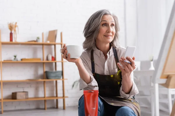 Smiling middle aged artist holding cup of coffee and smartphone near canvas — Stock Photo