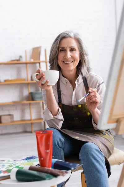 Happy middle aged artist holding cup of coffee and paintbrush near canvas — Stock Photo