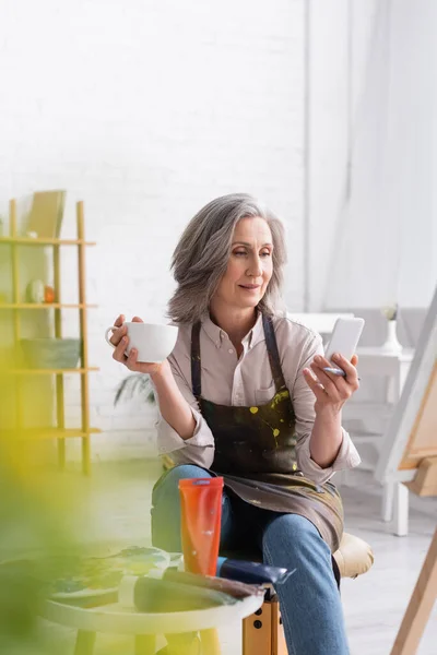 Artiste d'âge moyen tenant un pinceau, une tasse de café et utilisant un smartphone près de la toile et un premier plan flou — Photo de stock