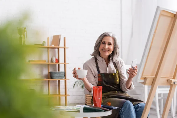 Heureux artiste d'âge moyen tenant pinceau, tasse de café et en utilisant smartphone près de la toile et au premier plan flou — Photo de stock