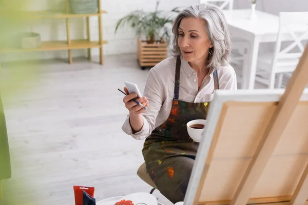 Middle aged artist holding paintbrush, cup of coffee and using smartphone near canvas — Stock Photo