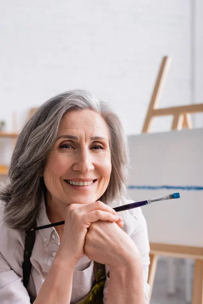 Joyful middle aged painter with grey hair holding paintbrush — Stock Photo