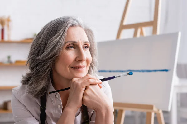 Happy middle aged painter with grey hair holding paintbrush while looking away — Stock Photo