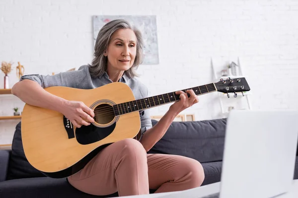 Жінка середнього віку з сірим волоссям, що навчається грати на акустичній гітарі біля ноутбука на розмитому передньому плані — стокове фото