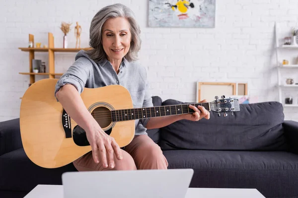 Жінка середнього віку з сірим волоссям, що навчається грати на акустичній гітарі, досягаючи ноутбука — стокове фото