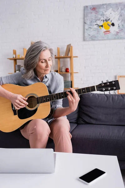 Mulher de meia idade aprendendo a tocar guitarra acústica perto de laptop e smartphone com tela em branco na mesa de café — Fotografia de Stock