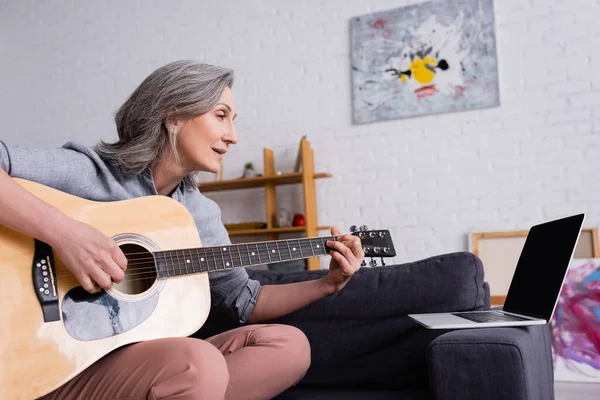 Mulher de meia-idade com cabelos grisalhos aprendendo a tocar guitarra acústica perto de laptop com tela em branco no sofá — Fotografia de Stock