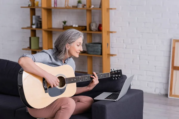 Happy middle aged woman with grey hair learning to play acoustic guitar near laptop on sofa — Stock Photo