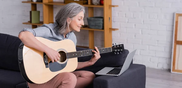 Happy middle aged woman with grey hair learning to play acoustic guitar near laptop on sofa, banner — Stock Photo