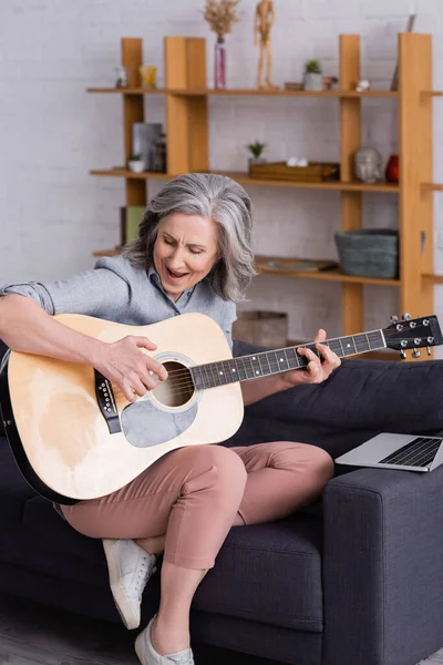 Middle aged woman with grey hair learning to play acoustic guitar while singing near laptop on sofa — Stock Photo