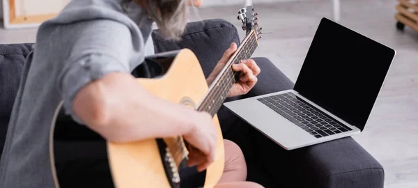 Обрізаний вид розмитої жінки середнього віку, яка вчиться грати на акустичній гітарі біля ноутбука з порожнім екраном на дивані, банер — стокове фото