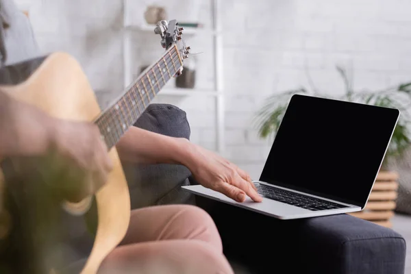 Vista recortada de mujer de mediana edad borrosa aprendiendo a tocar la guitarra acústica cerca de la computadora portátil con pantalla en blanco en el sofá - foto de stock