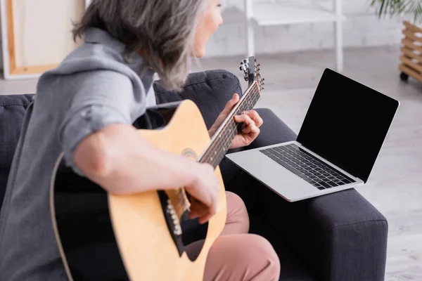Femme d'âge moyen floue apprenant à jouer de la guitare acoustique près d'un ordinateur portable avec écran blanc sur le canapé — Photo de stock