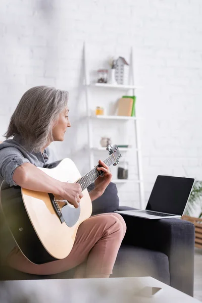 Женщина средних лет учится играть на акустической гитаре рядом с ноутбуком с пустым экраном на диване — стоковое фото