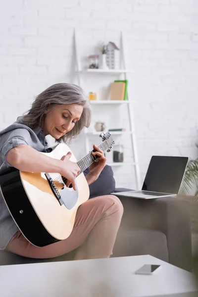 Femme mûre apprenant à jouer de la guitare acoustique près d'un ordinateur portable avec écran vide sur le canapé — Photo de stock