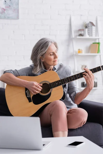 Mujer madura aprendiendo a tocar la guitarra acústica cerca del ordenador portátil y el teléfono inteligente con pantalla en blanco - foto de stock