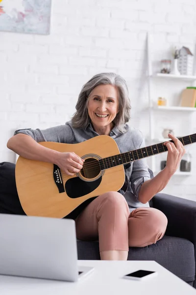 Happy mature woman learning to play acoustic guitar near laptop and smartphone with blank screen — Stock Photo