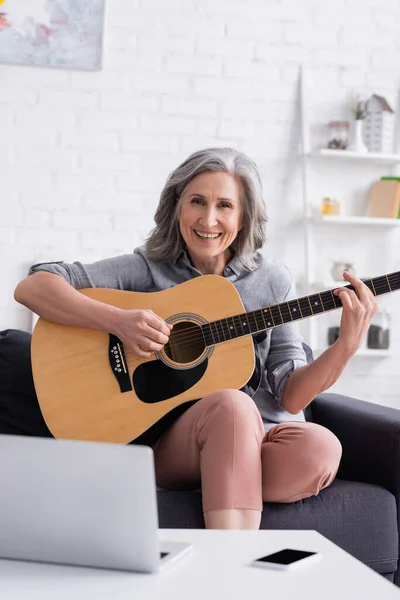 Mulher madura feliz aprendendo a tocar guitarra acústica perto de laptop e smartphone com tela em branco — Fotografia de Stock