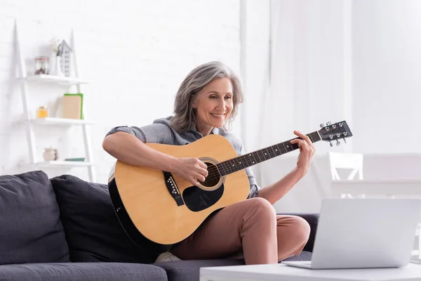 Щаслива жінка середнього віку, яка вчиться грати на акустичній гітарі біля ноутбука на кавовому столі — стокове фото