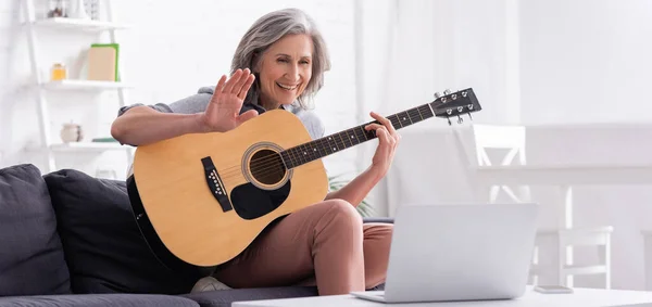 Mulher de meia-idade com cabelos grisalhos segurando guitarra acústica enquanto acena a mão durante videochamada no laptop, banner — Fotografia de Stock