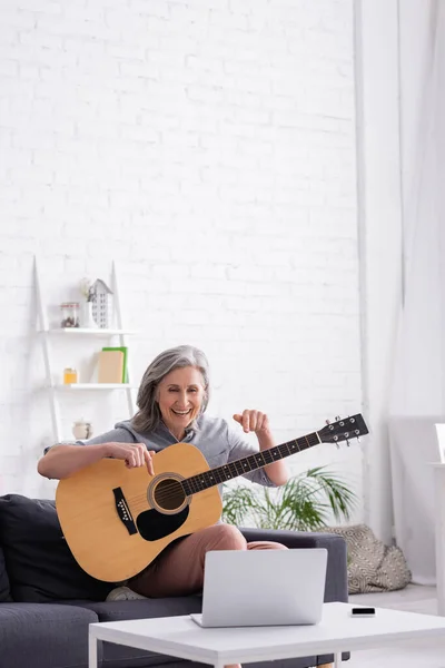 Middle aged woman with grey hair pointing at acoustic guitar during video call on laptop — Stock Photo