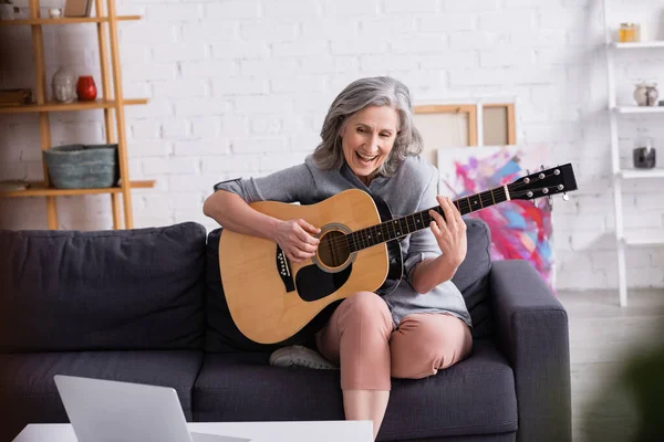 Mulher de meia-idade espantada com cabelos grisalhos aprendendo a tocar guitarra acústica perto do laptop — Fotografia de Stock