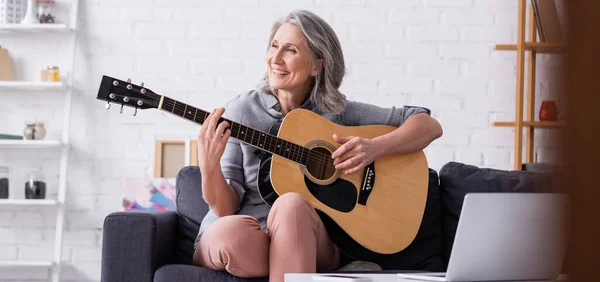 Задоволена зріла жінка з сірим волоссям вчиться грати на акустичній гітарі біля ноутбука, банер — стокове фото