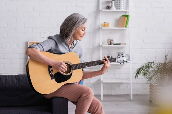 Mujer madura con pelo gris tocando la guitarra acústica en la sala de estar - foto de stock
