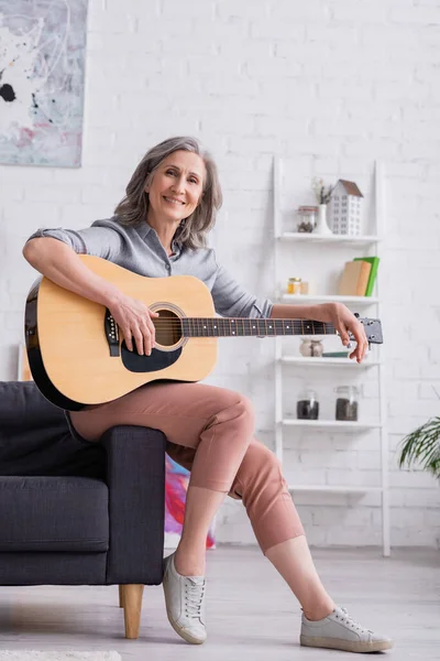 Улыбающаяся зрелая женщина с седыми волосами, сидящая с акустической гитарой в гостиной — стоковое фото