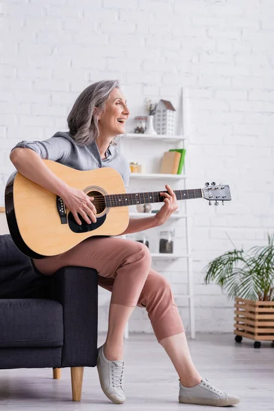 Mujer madura alegre con pelo gris tocando la guitarra acústica mientras está sentado en la sala de estar - foto de stock