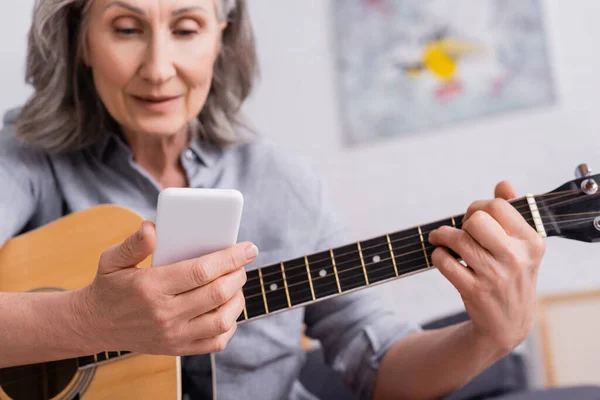 Femme mature floue avec les cheveux gris tenant smartphone tout en apprenant à jouer de la guitare acoustique — Photo de stock