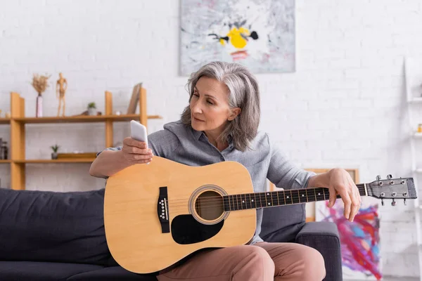 Mujer madura con pelo gris mirando el teléfono inteligente mientras aprende a tocar la guitarra acústica - foto de stock